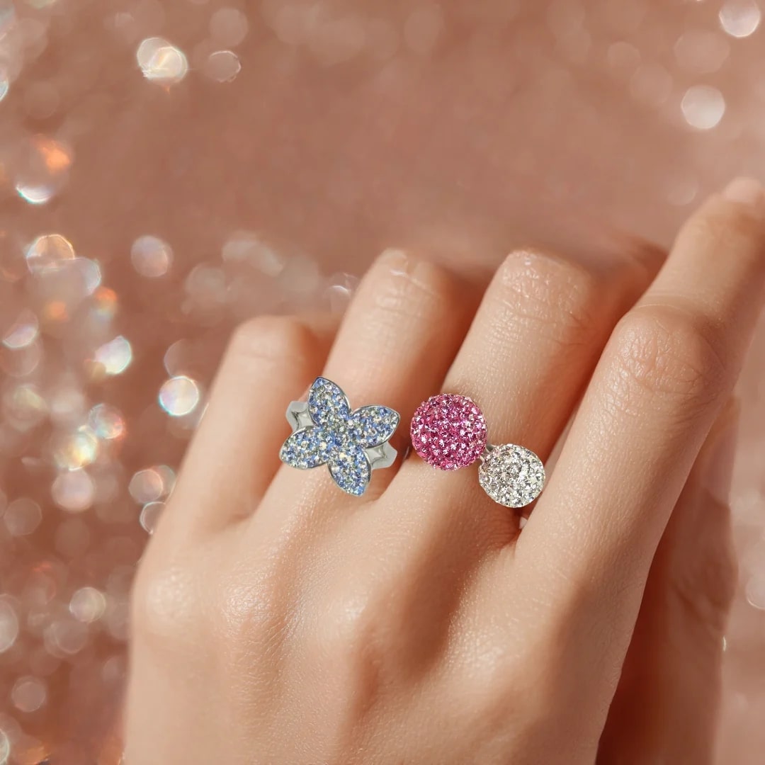 Close-up of a hand wearing three sparkling rings on one finger: a blue flower/butterfly ring, and two interconnected spherical rings, one pink and one clear/white in shiny pinkish background